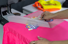 A person carefully applying a colorful DTF transfer design onto a bright pink t-shirt using a heat press machine.